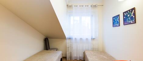 Una habitación pequeña y luminosa con dos camas individuales, suelo de madera, una ventana con cortinas translúcidas y dos cuadros enmarcados en la pared.