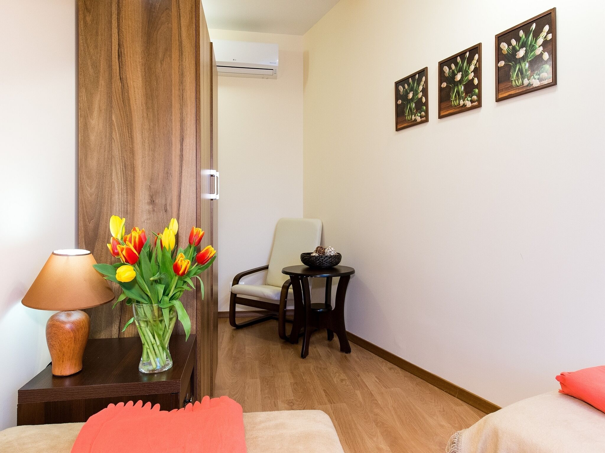 Une chambre d’hôtel avec un plancher en bois, un bureau avec une lampe et un vase de tulipes, une chaise et un lit avec un oreiller rouge.