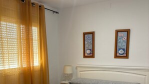 A neatly made bed with a grey patterned bedspread and two towels folded on it. There is a nightstand with a lamp beside the bed, and two framed pictures on the wall. - ISLANTUR BELLAMAR (Lepe)
