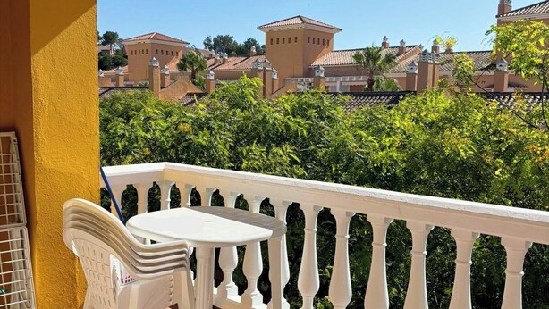 A balcony with a white chair and table, a yellow wall, and a view of buildings and greenery. - ISLANTUR BELLAMAR (Lepe)