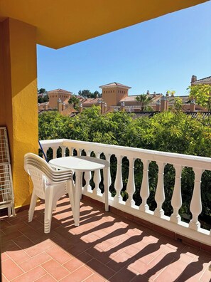 Un balcon avec une chaise et une table blanches, un mur jaune et une vue sur des bâtiments et de la verdure.