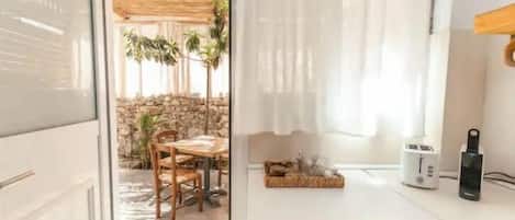 Cocina compacta con encimera blanca, tostadora y cafetera. Zona de comedor con mesa y sillas, y vista a un muro de piedra y plantas al exterior.