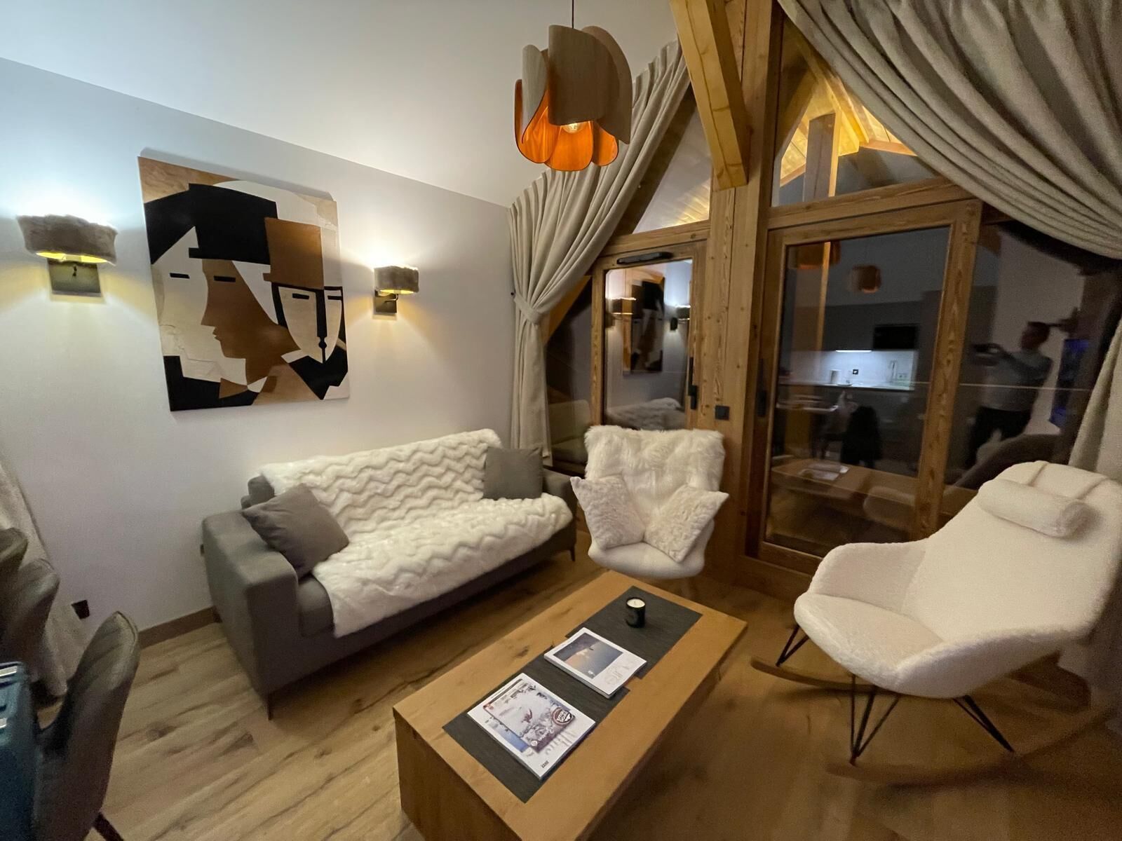 A modern living room with a sofa, armchair, and coffee table, wooden flooring, and a large window with curtains.