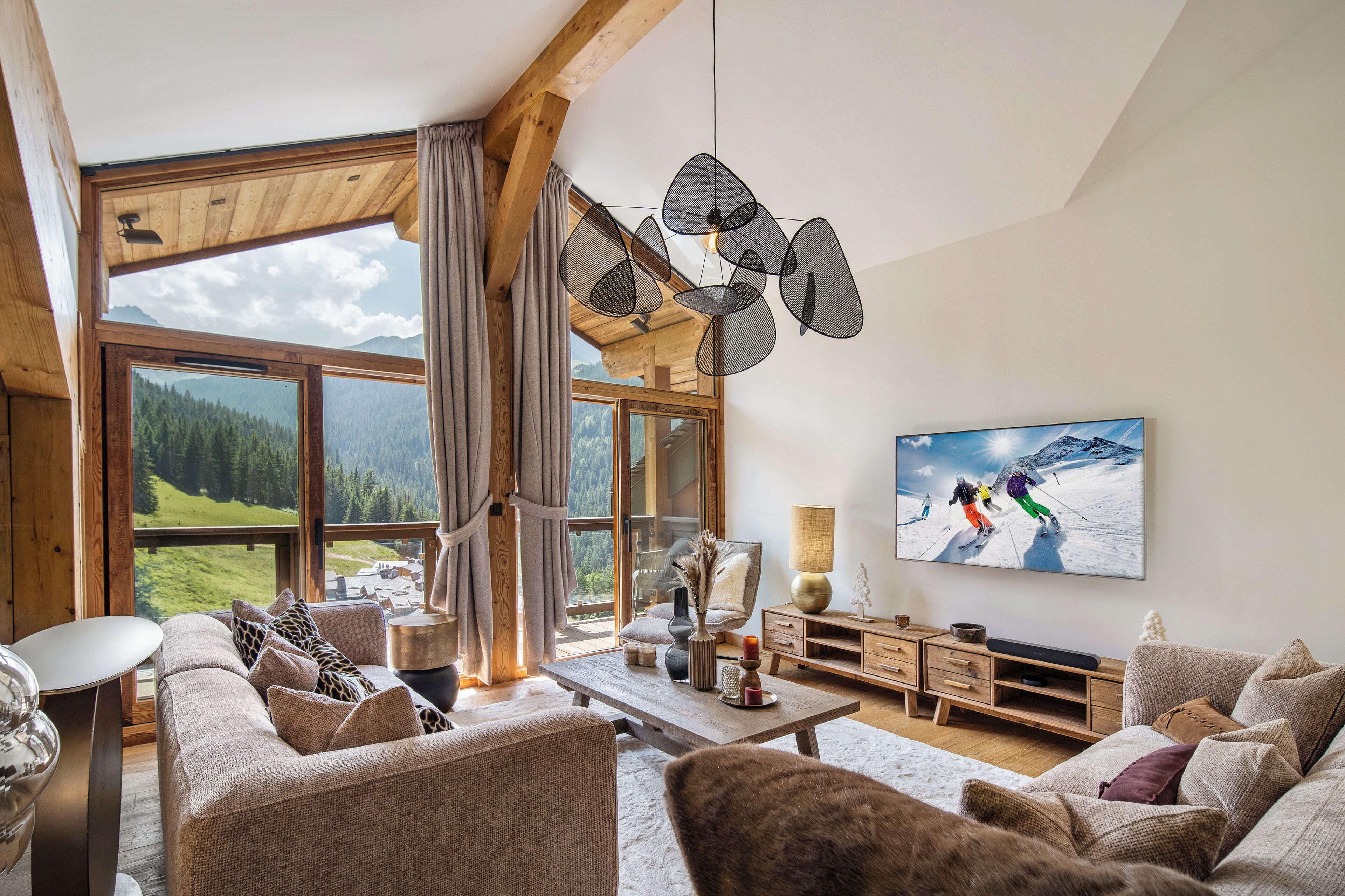 A spacious living room with a large window, wooden beams, a modern chandelier, and a television displaying a skiing scene.
