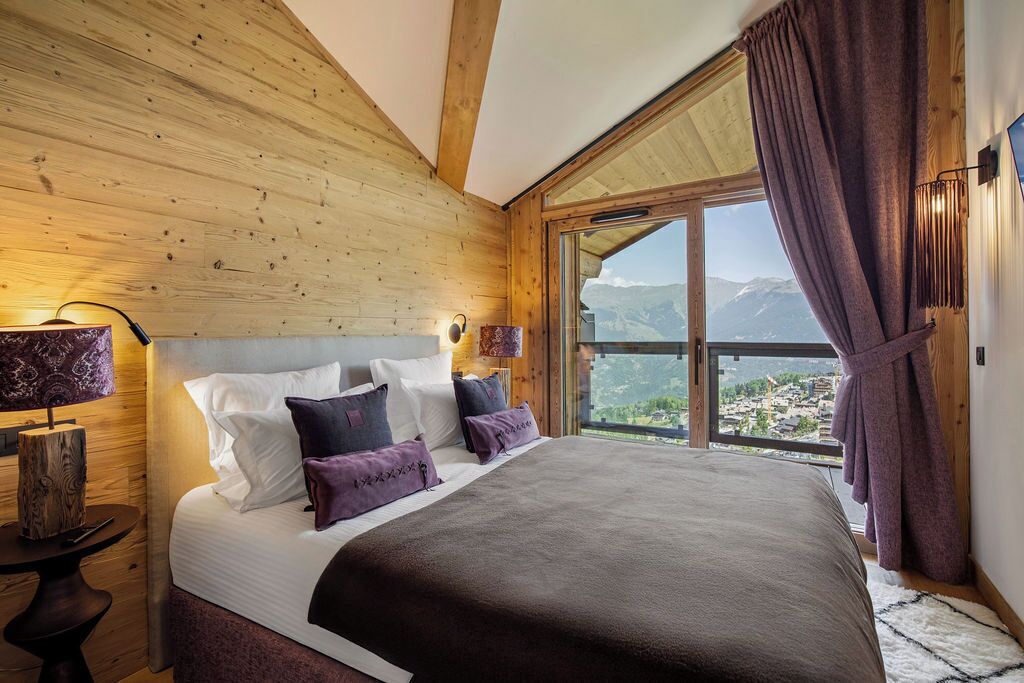 Quarto com uma cama grande, paredes de madeira, vista para montanhas e uma janela com cortinas.