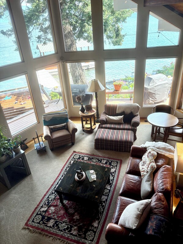 A spacious living room with large windows, a fireplace, a coffee table, and a view of the water.