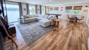 A spacious living room with a large sectional sofa, a dining table with chairs, and a view of the outdoors through large windows. - The Belmont Lake Retreat -Sleeps 14! (Belmont)