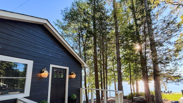 A dark wooden house with a white porch and railing, surrounded by trees and a body of water. - The Belmont Lake Retreat -Sleeps 14! (Belmont)