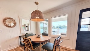 A dining area with a round wooden table, four chairs, a hanging light fixture, and large windows offering a view of the outdoors. - The Belmont Lake Retreat -Sleeps 14! (Belmont)