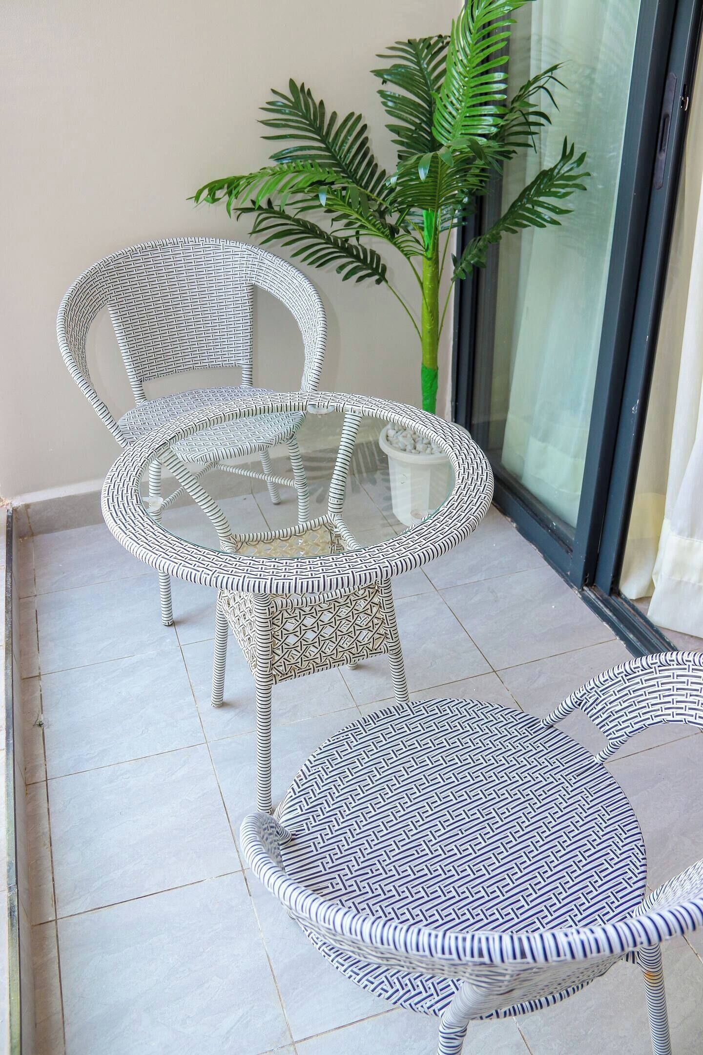 A balcony with two wicker chairs and a glass table, a potted plant, and a sliding glass door.