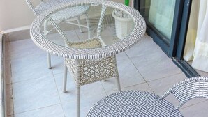 A balcony with two wicker chairs and a glass table, a potted plant, and a sliding glass door.