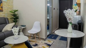 A modern living room with a grey sofa, a white chair, a small white table, and a white vase with flowers. There is a plant on the left and a door in the background.