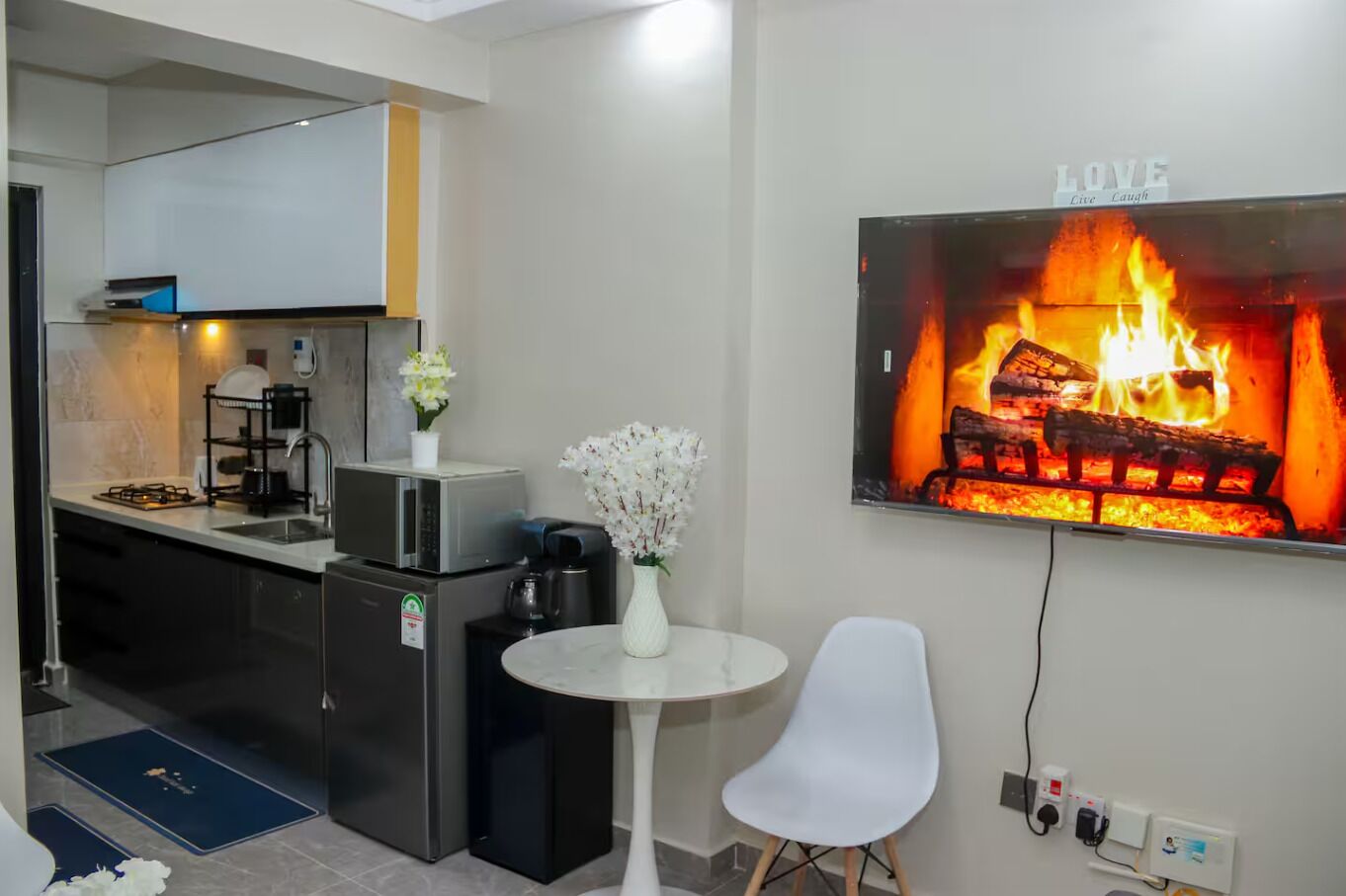 A modern kitchen with a microwave, stove, and coffee maker. There is a white chair and a small table with a vase of flowers. A fireplace with a 'LOVE' sign is on the wall.