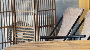 Ein Zimmer mit einem Holztisch, zwei Korbstühlen, einer Metallsofa und einem Holztrenner mit vertikalen Lamellen.