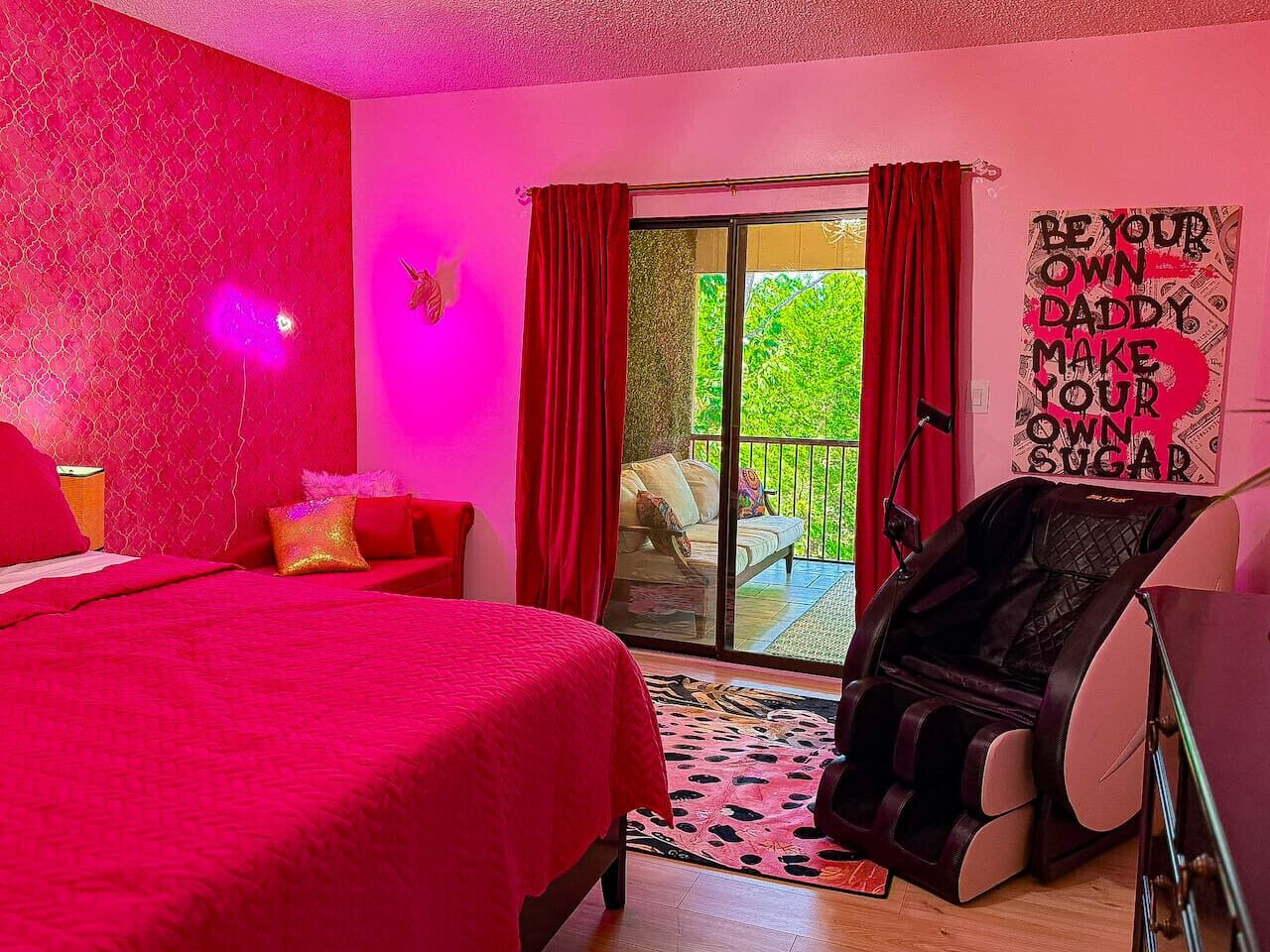 A bedroom with a red bedspread, a massage chair, and a view of a balcony with red curtains.