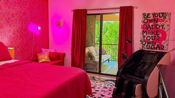 A bedroom with a red bedspread, a massage chair, and a view of a balcony with red curtains.