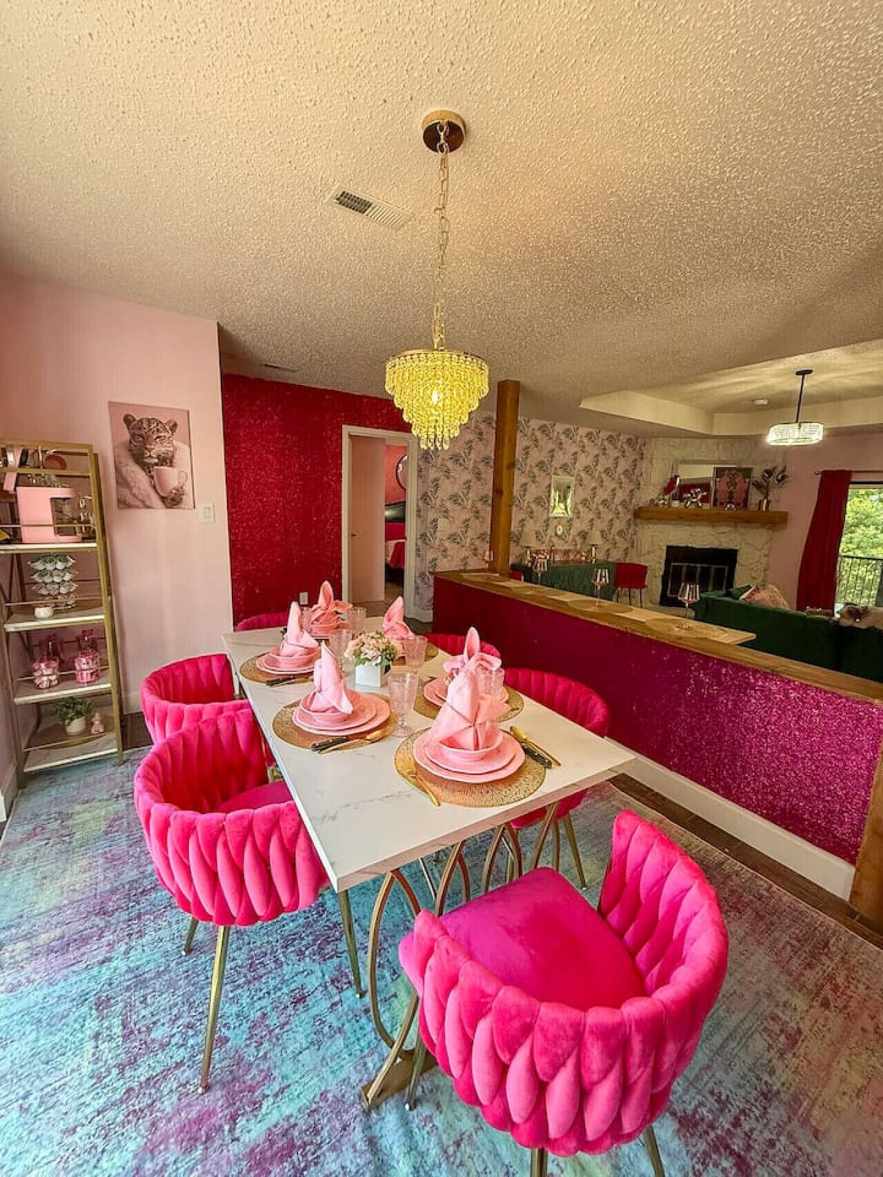 Un comedor con una mesa blanca dispuesta para cuatro comensales, sillas tapizadas en rosa, una araña y un estampado rojo y blanco en la pared.