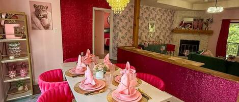 Un comedor con una mesa blanca dispuesta para cuatro comensales, sillas tapizadas en rosa, una araña y un estampado rojo y blanco en la pared.