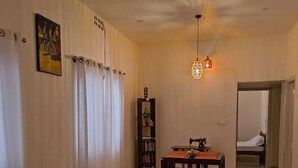 A living room with a sofa, armchair, and a small table with a plant. There is a bookshelf and a sewing machine on a table. The room has white curtains and a ceiling light fixture. - Serene 3-bedroom house in Mysore (Mysore)