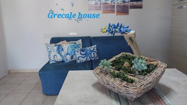 Quarto com um sofá azul, almofadas decorativas, uma parede com o letreiro 'Grecale house' e um quadro na parede retratando uma cena de praia.