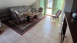 A cozy living room with a high ceiling, a wooden beam, a dining table with chairs, a patterned rug, and a view of the outdoors through a window with green curtains.
