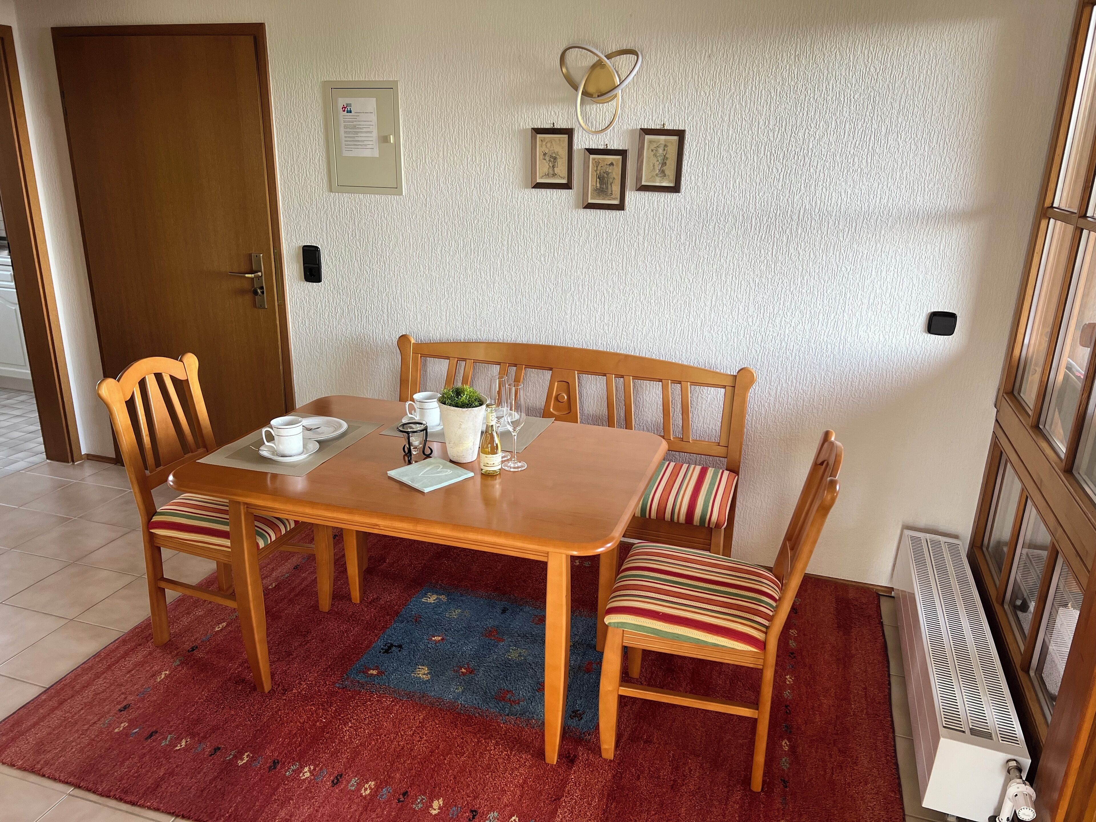 A dining area with a wooden table, chairs, a bench, a red rug, a potted plant, and framed pictures on the wall.