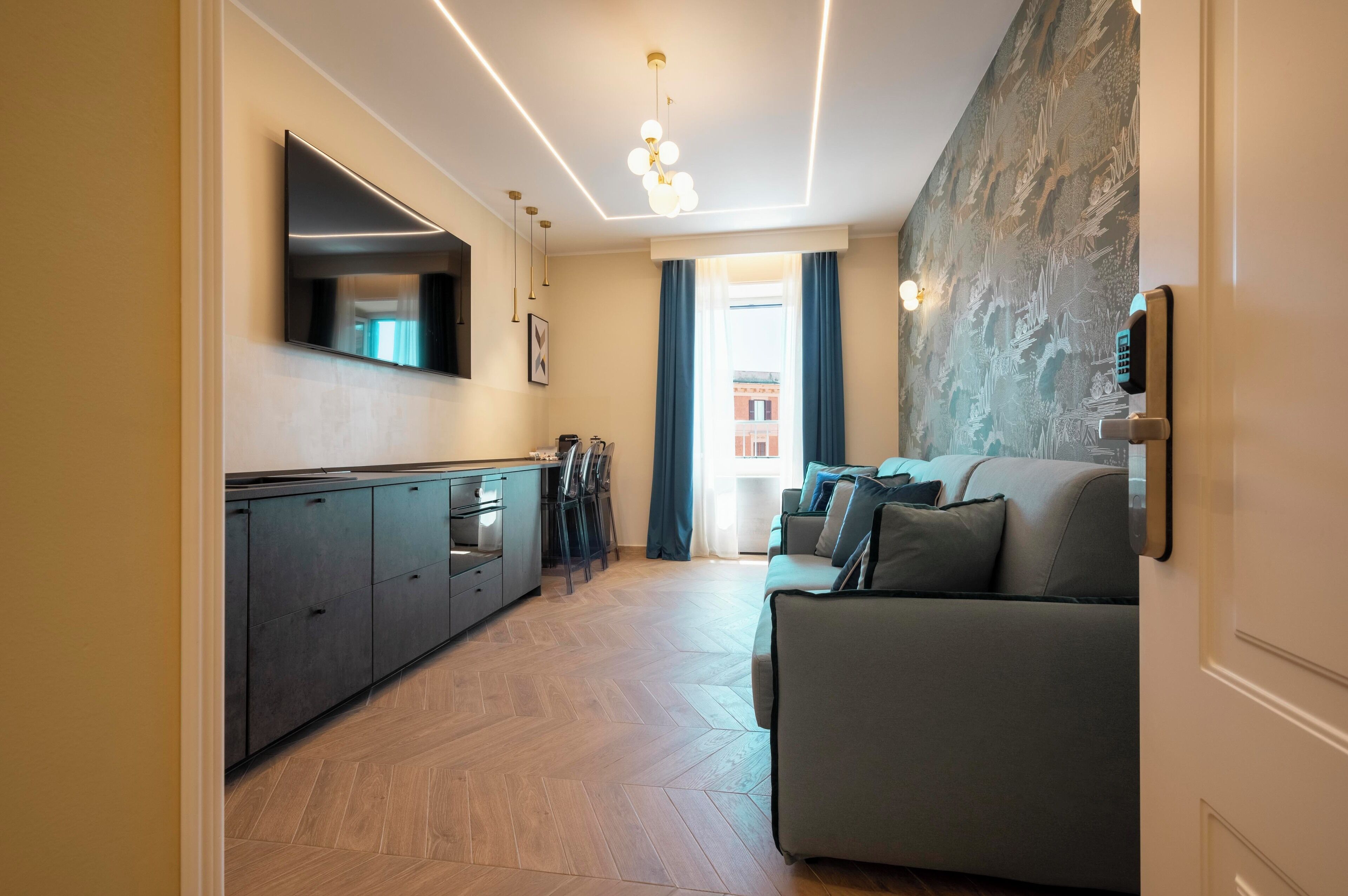 A modern living room with a sofa, a flat-screen TV mounted on the wall, a kitchen area with cabinets and a countertop, and a patterned wall.