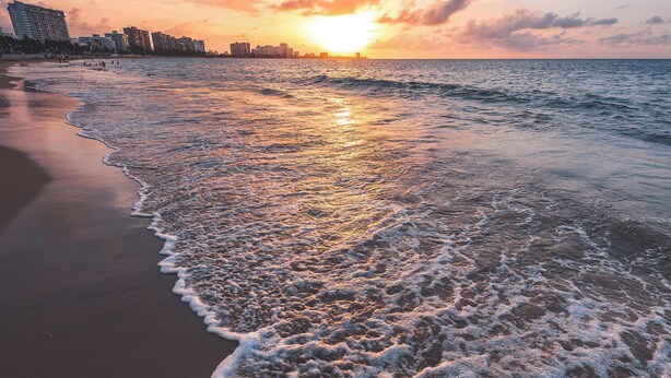 A beach with waves at sunset, a city skyline in the background, and a clear sky.
