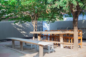 Un espacio al aire libre con bancos y mesas de madera, rodeado de árboles y un muro bajo.