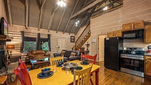A log cabin interior with a dining area, a living room with a leather sofa, a kitchen with modern appliances, and a wooden staircase.