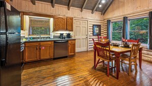 A log cabin kitchen with wooden cabinets, a dining table with chairs, and a large window with curtains.