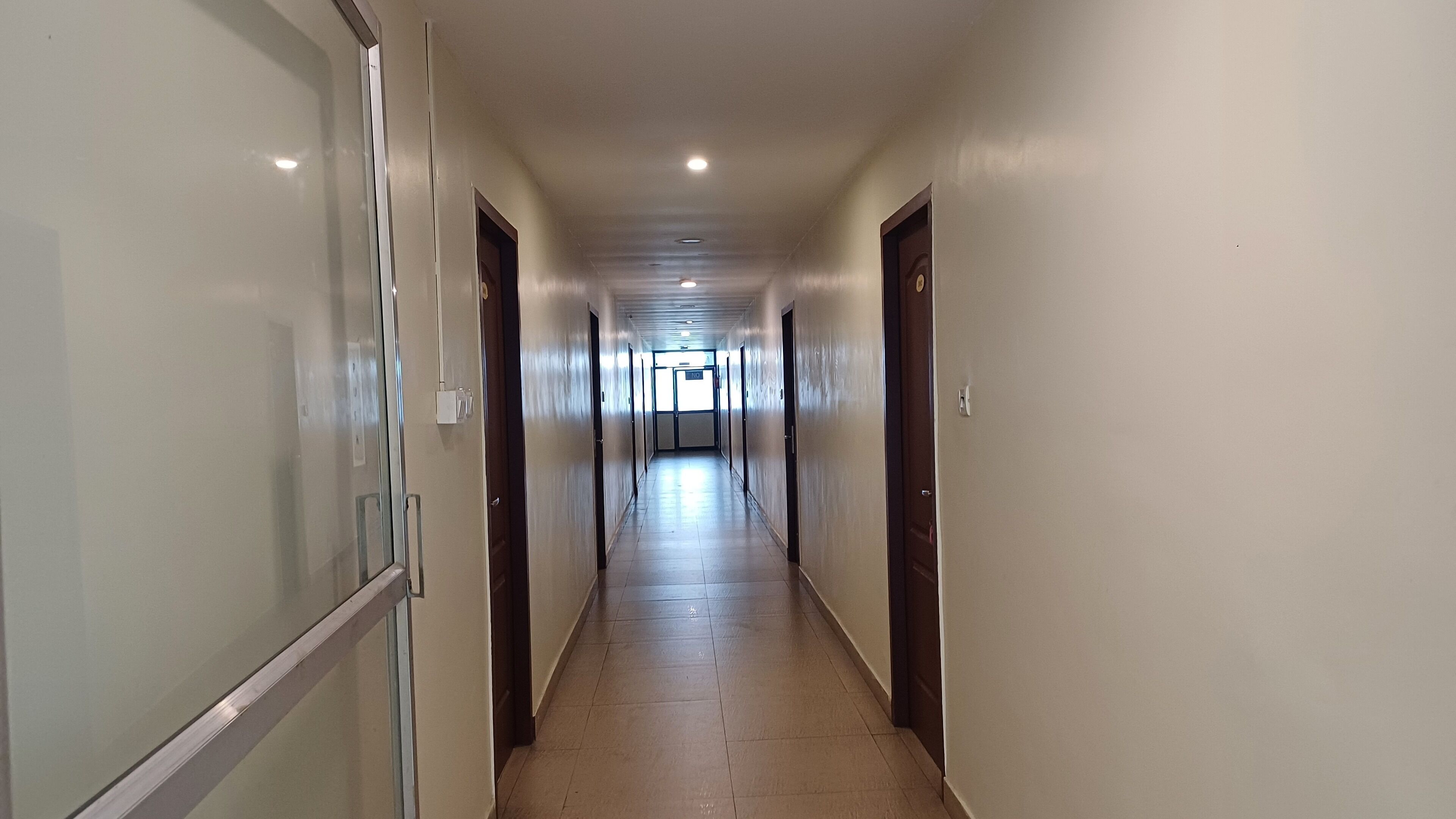 A long, narrow hallway with doors on both sides leading to rooms. The hallway has a tiled floor and is well-lit with ceiling lights.
