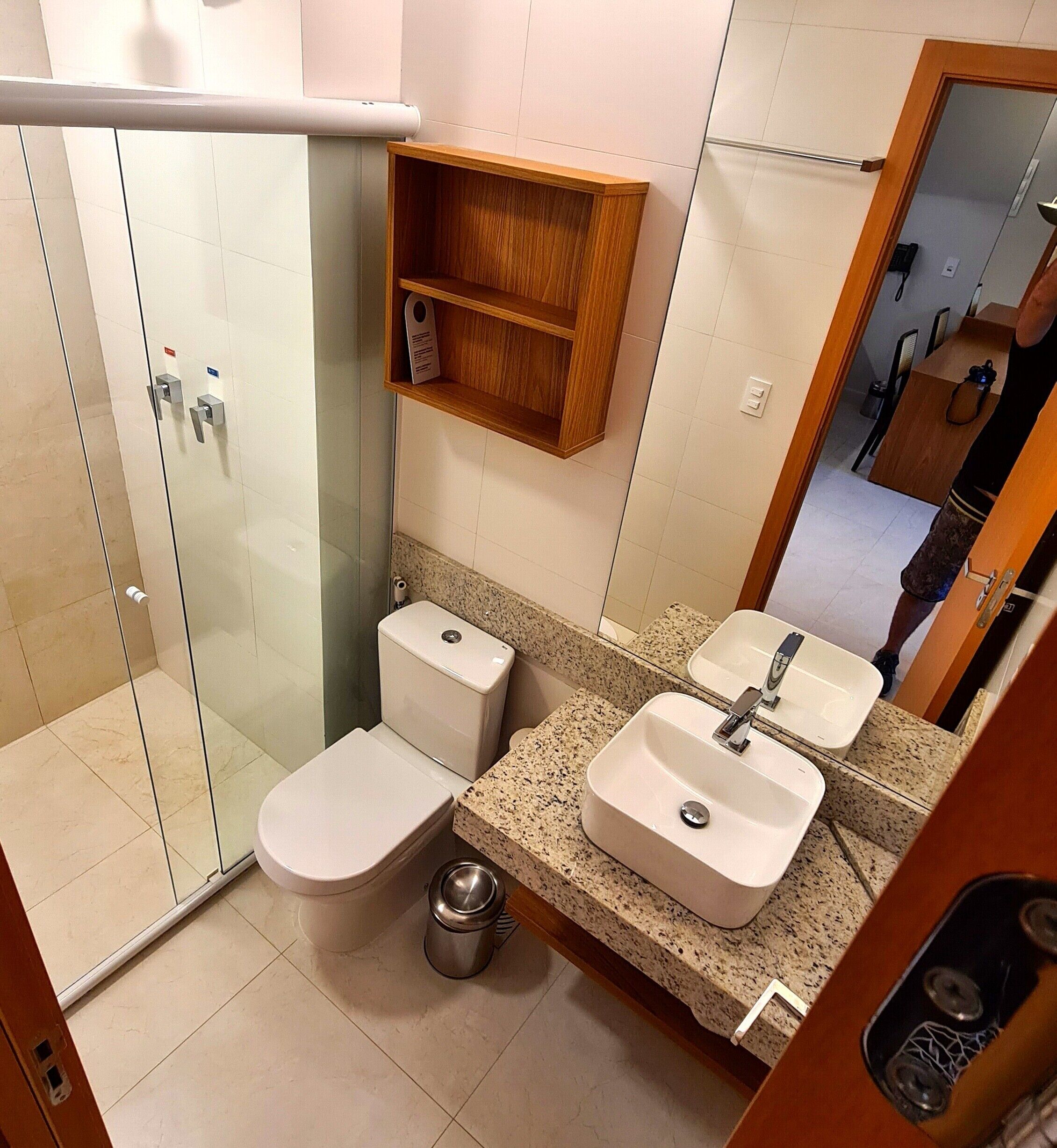 Un baño con ducha de vidrio, inodoro, lavamanos y estante de madera sobre el lavamanos.