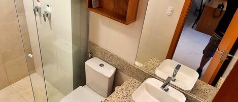 Un baño con ducha de vidrio, inodoro, lavamanos y estante de madera sobre el lavamanos.