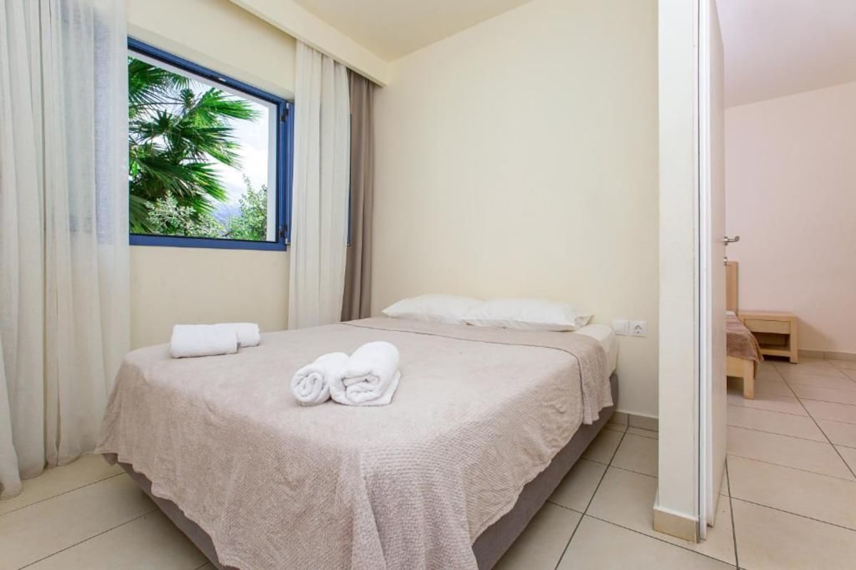 A neatly made bed with a beige bedspread and two rolled towels, a window with a view of palm trees, and a wooden bench in the corner.