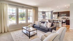 A spacious living room with a grey sofa, patterned armchairs, and a coffee table. A kitchen with dark cabinets and a microwave is visible in the background. - 4 Mi to The District: Family Home w/ Tennis Access (South Jordan)