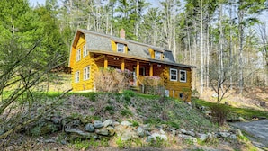 Une cabane en rondins dotée d’un porche, entourée d’arbres et d’un paysage rocheux.