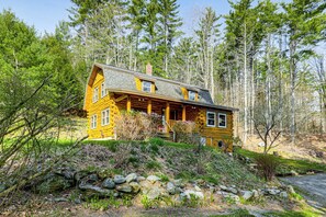 A log cabin with a porch, surrounded by trees and a rocky hill. - Forest Hideaway in Plainfield: Hike, Paddle & Ski (Plainfield)