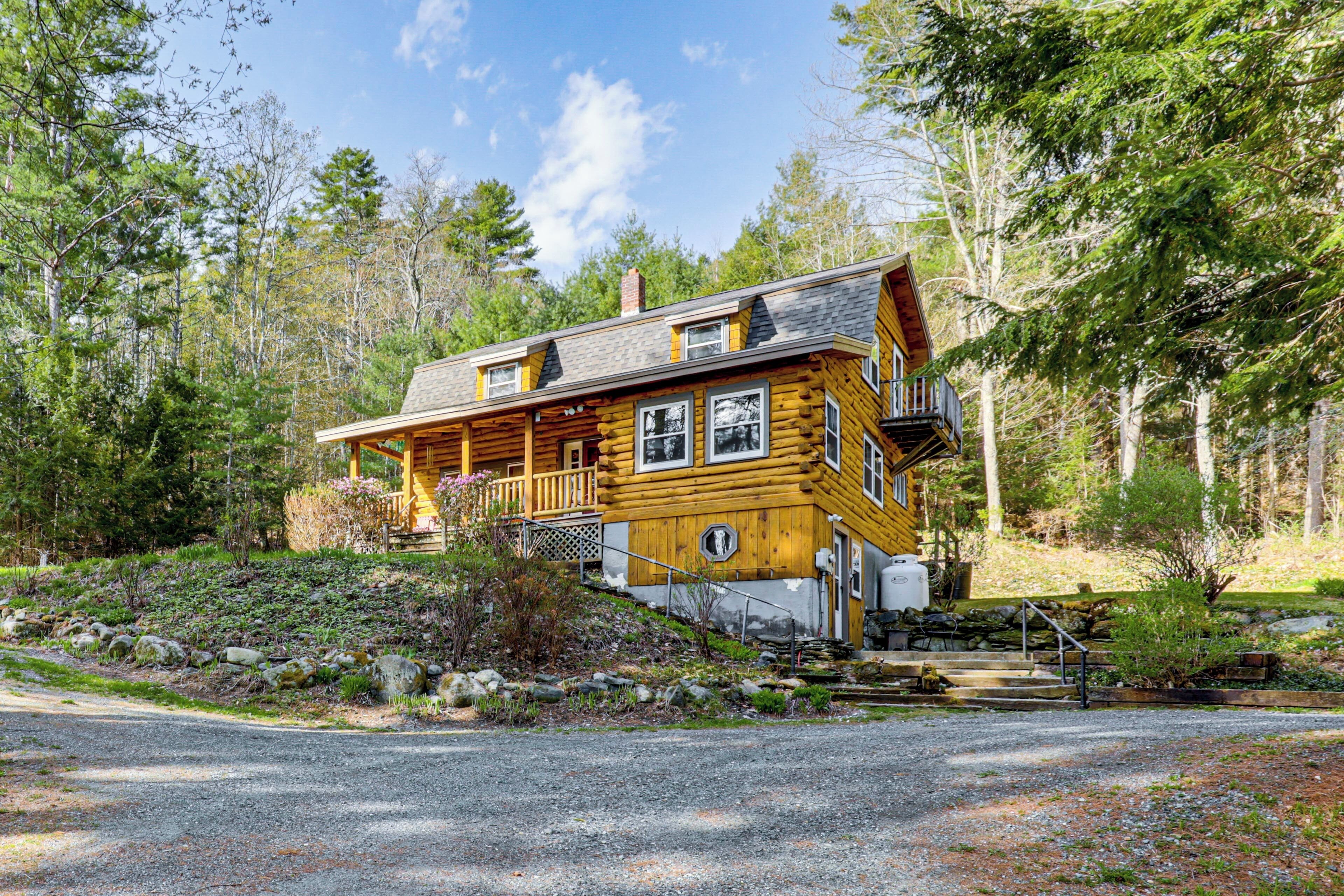 Une cabane en rondins avec un porche, entourée d’arbres et bordée d’une allée de gravier.