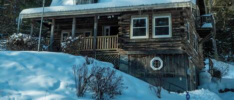 Une cabane en rondins dotée d’un porche et d’une terrasse supérieure couverte, entourée de neige et d’arbres.