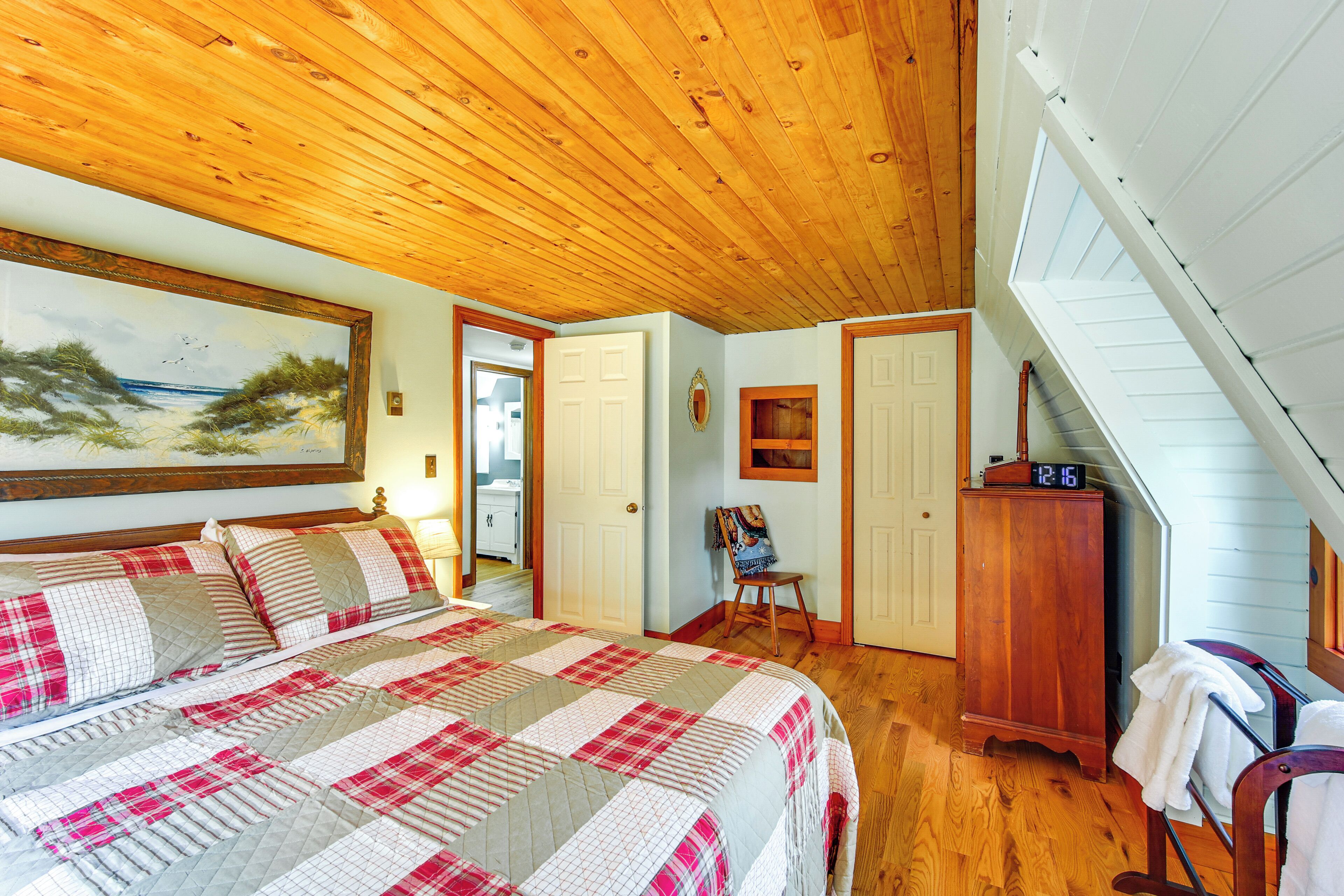 Une chambre avec un plafond en bois, un lit recouvert d’une couverture à carreaux, un tableau accroché au mur et une horloge posée sur une commode.