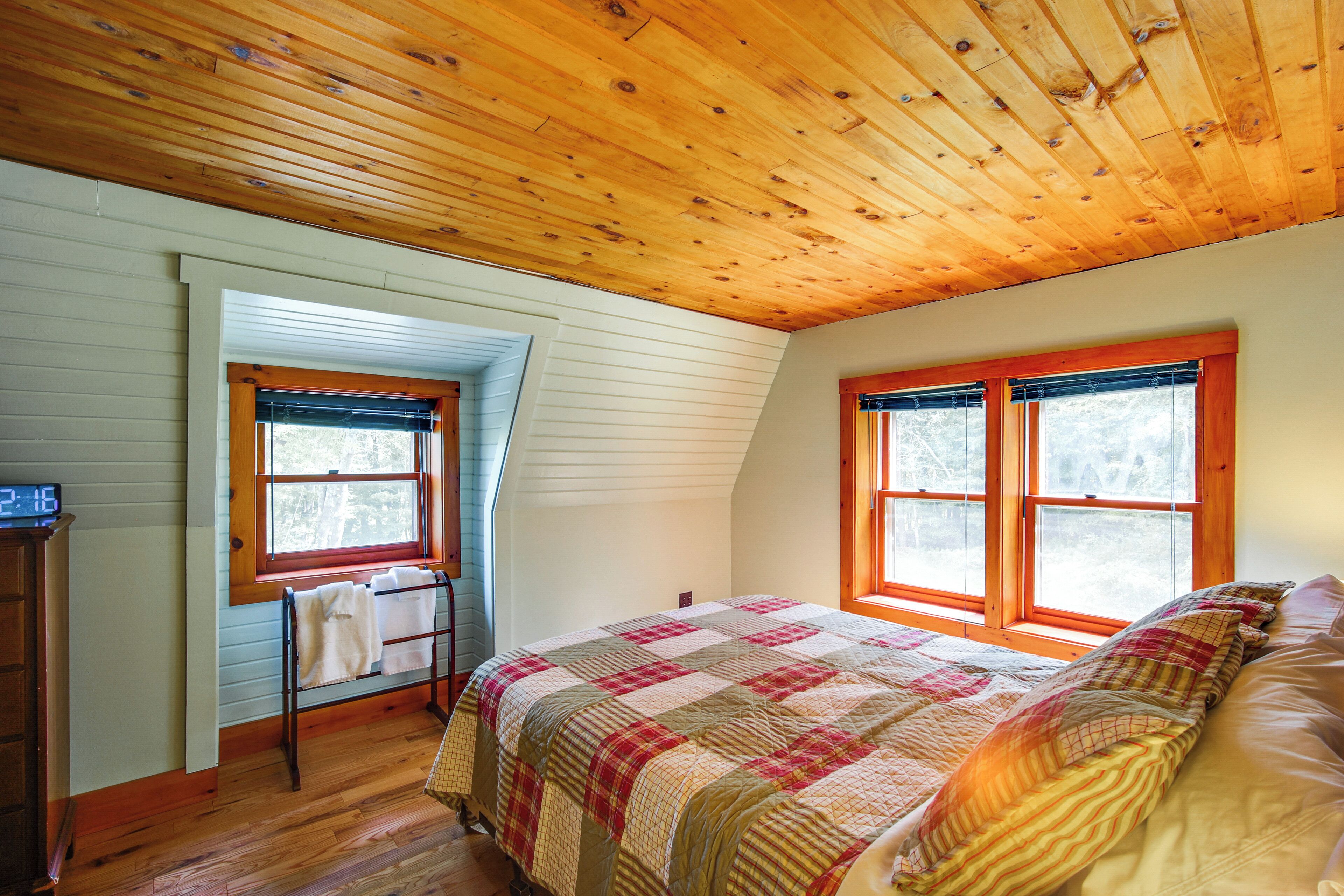 A bedroom with a wooden ceiling, a bed with a quilt, two windows, and a nightstand with a clock.