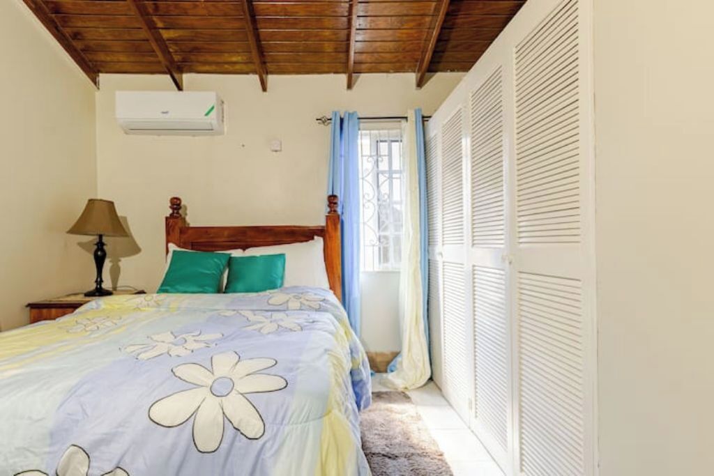A bedroom with a wooden ceiling, a bed with floral bedding, a nightstand with a lamp, and a window with curtains.