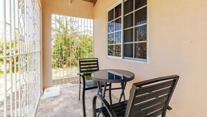 Une terrasse couverte avec une table ronde et deux chaises, une fenêtre donnant sur un paysage verdoyant et un portail en métal blanc.