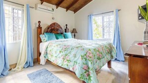 Une chambre avec un plafond en bois, un lit recouvert d’une literie à motifs floraux, une tête de lit en bois et une fenêtre avec des rideaux.