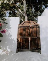 Una verja de madera con un pestillo de metal, frente a una pared blanca con un árbol y flores rosadas a la izquierda.