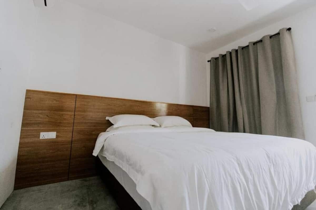 Habitación individual con una cama de madera, ropa de cama blanca y una cortina gris.