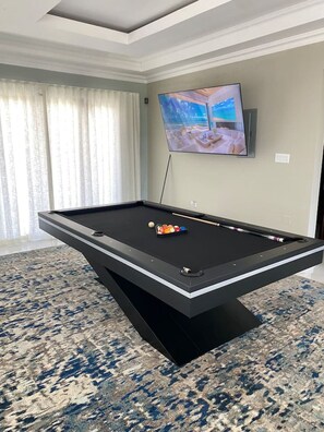 A pool table with balls, a television mounted on the wall, and a window with curtains.