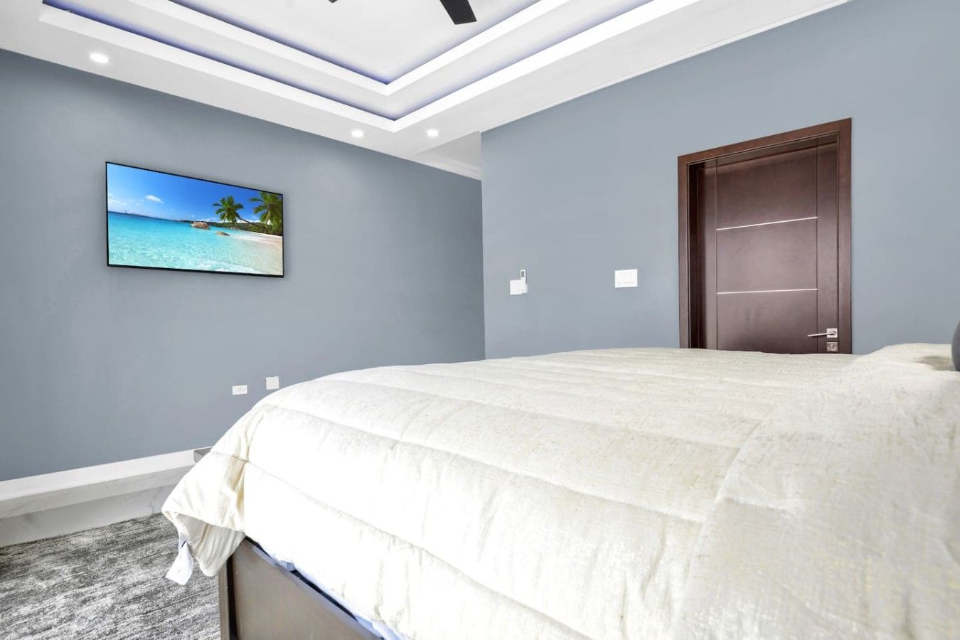 Een slaapkamer met een groot bed, een deur, een aan de muur bevestigde televisie en een plafond met inbouwverlichting.