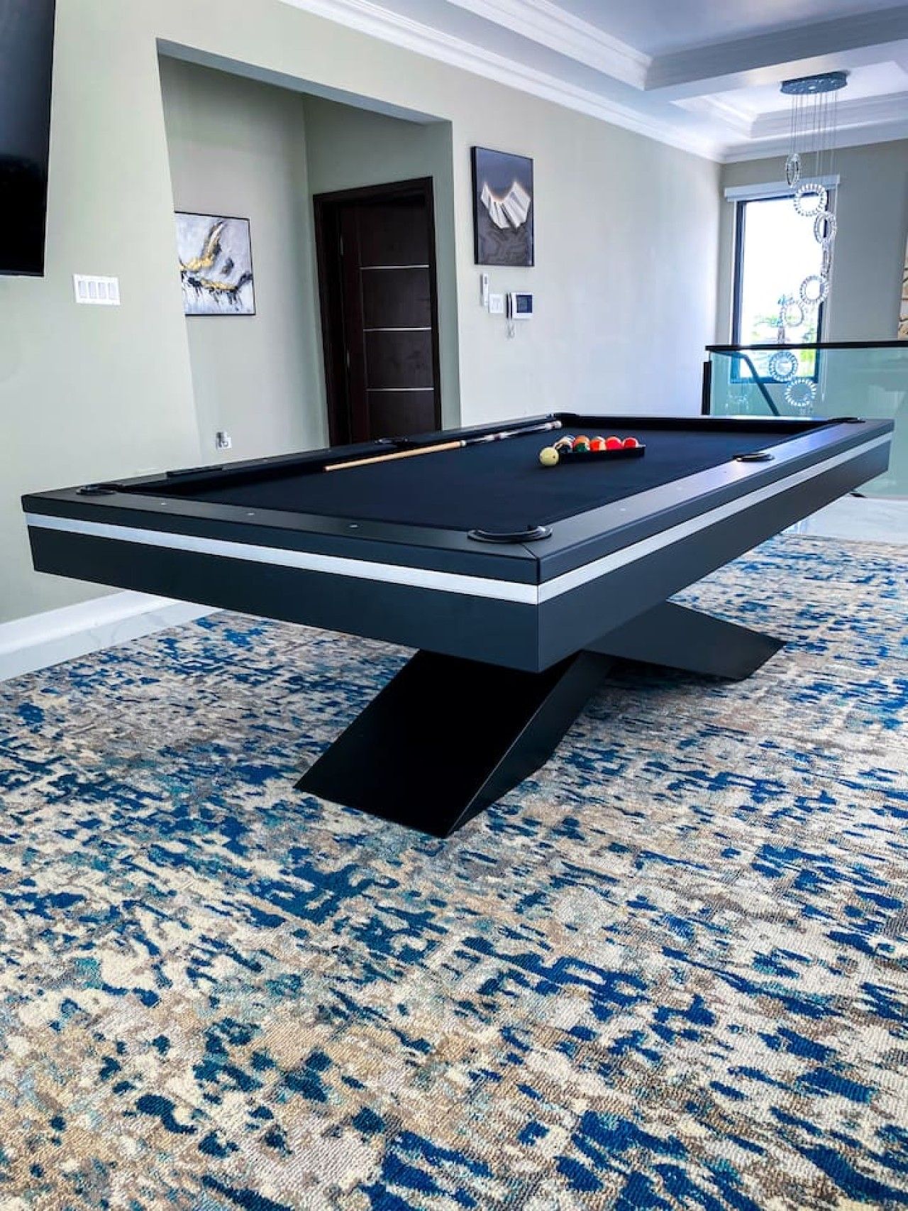 Une table de billard dans une pièce dotée d’un tapis à motifs, d’une porte et d’œuvres d’art accrochées aux murs.